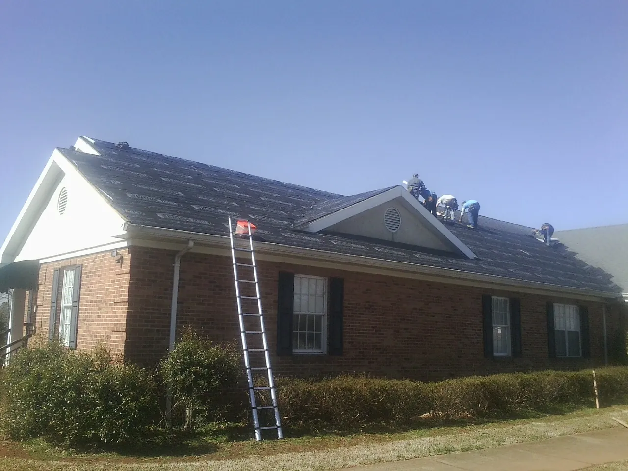 Crew tearing shingles off a brick ranch roof with a ladder set — Emergency Roof Repair work by our crew in Greensboro